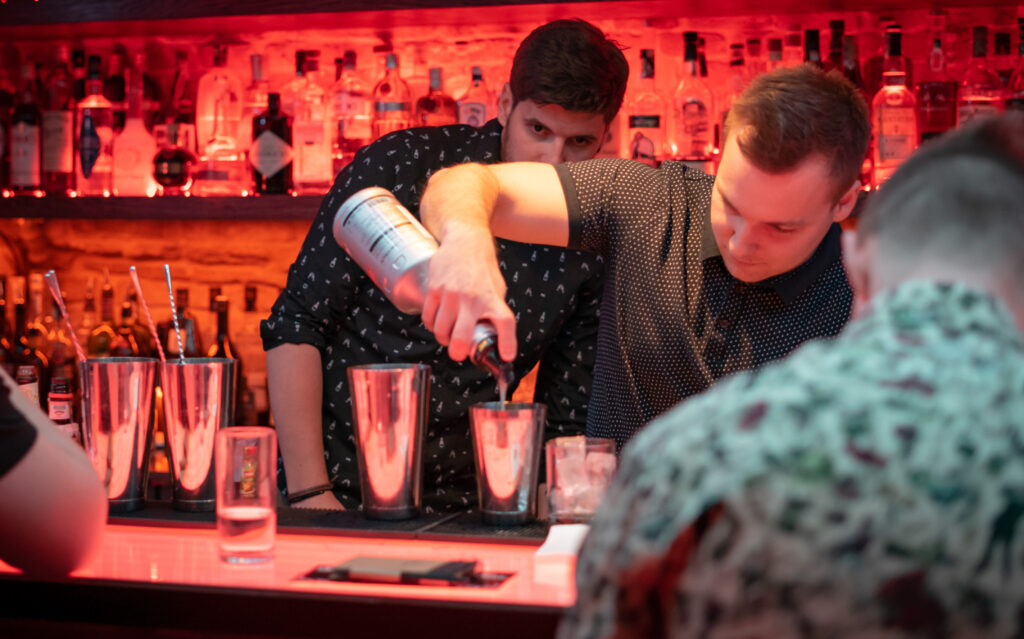 Participant of the barman class tries to pour alcohol into several glasses while the trainer observes him