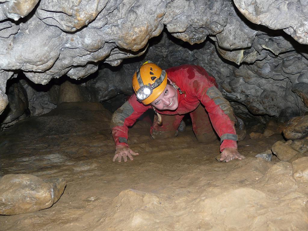 A lady in a red jumpsuit with a yellow helmet crawls around the cave