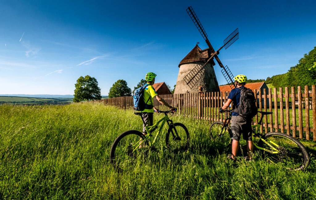 Two cyclists standing in the tall grass in the meadow behind the fence where the windmill is located