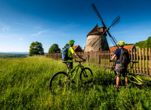 Two cyclists standing in the tall grass in the meadow behind the fence where the windmill is located