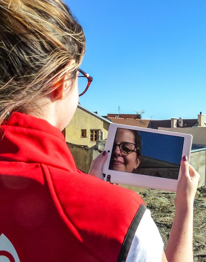 A woman in red vest standing with her back to the photographer, holding a tablet in which her reflection can be seen